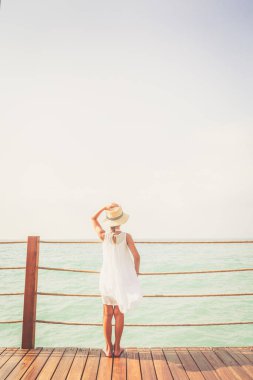 Beyaz elbiseli bronzlaşmış kız ahşap iskelede deniz manzarasının tadını çıkarıyor.