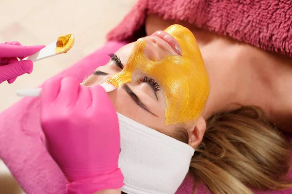 Cosmetic mask. The beautician applies a golden mask to a woman\'s face. Beautiful woman in a beauty salon during facial treatment