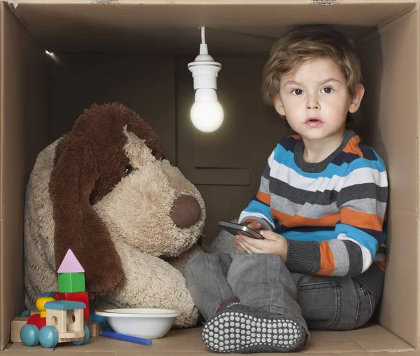 Niño lindo con su juguete relleno en caja de cartón con bombilla desnuda, que representa un pequeño lugar estrecho — Foto de Stock