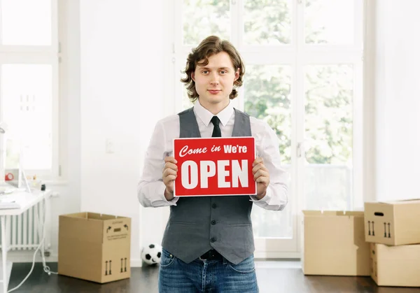 Young Business men Holding up a shop sign saying: Come in, We\'re Open. Surrounded by Moving boxes Start-up Business Concept