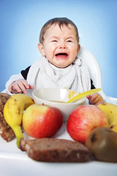 Crying unhappy Baby boy must eat healthy food for breakfast on bright blue background — Stock Photo, Image