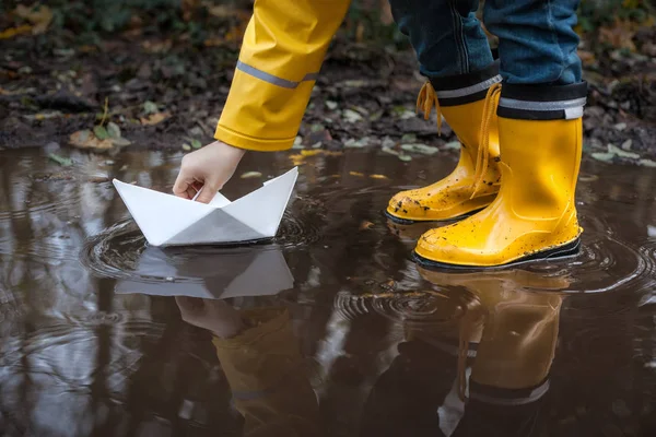 Παιδί με κίτρινη βροχή μπότες και μια μικρή λευκή βάρκα / πλοίου: παίζοντας σε μια λακκούβα, φαντάζονται τις περιπέτειές του — Φωτογραφία Αρχείου