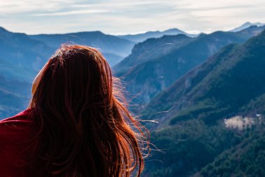Güneşli bir günde yüzü olmayan, dağ vadisi manzarasının önünde saçları dalgalanan kızıl saçlı genç bir kadının yakın plan görüntüsü.