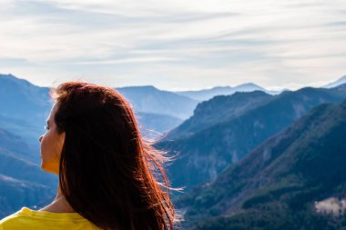 Güneşli bir günde, dağ vadisi manzarasının önünde dalgalanan uzun saçlı kızıl saçlı genç bir kadının yakın plan görüntüsü.
