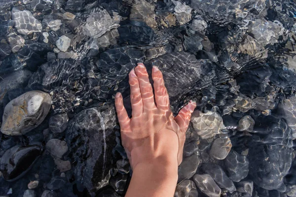 A young woman\'s hand underwater touching pebbles under clean transparent water of a mountain river with sunlight reflecting and flickering