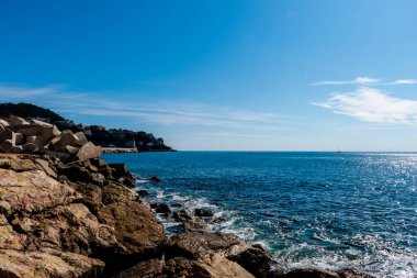 Akdeniz dalgaları kıyıdaki büyük kayalara ulaşırken, turkuaz suya yansıyan güneş ışığı ve güneşli bir günde arka planda bir yarımada (Güzel, Fransa)