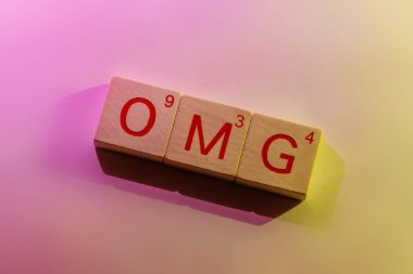 'omg' harfini heceleyen ahşap kare karoların yakın plan bir görüntüsü. Yumuşak pembe / mor ve zıt taraflardan gelen sarı / altın ışıkla aydınlatılıyor.