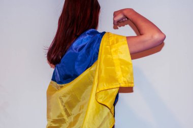 Arka planda, Ukrayna ulusal bayrağı giymiş, yumruğunu sıkarak gücünü gösteren bir pelerin giymiş, tanınamayan genç, beyaz saçlı bir kadının yakın plan fotoğrafı.