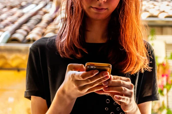 A close-up shot of an unrecognizable young Caucasian redhead woman looking down at her smartphone with a neutral facial expression with an old French building in the background