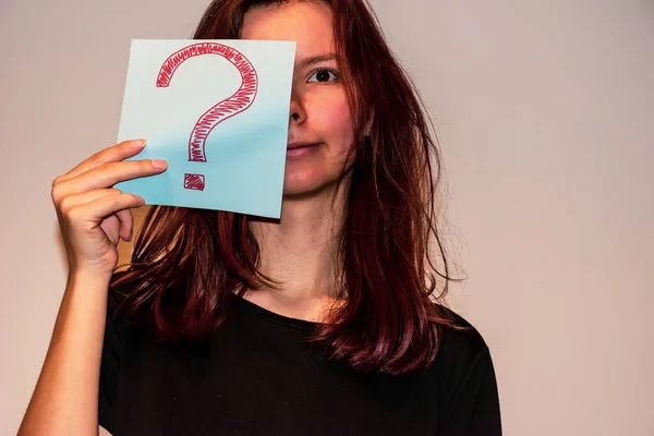 A close-up shot of a young Caucasian redhead woman hiding a half of her face behind a light blue paper sheet with a hand-drawn question mark and smiling