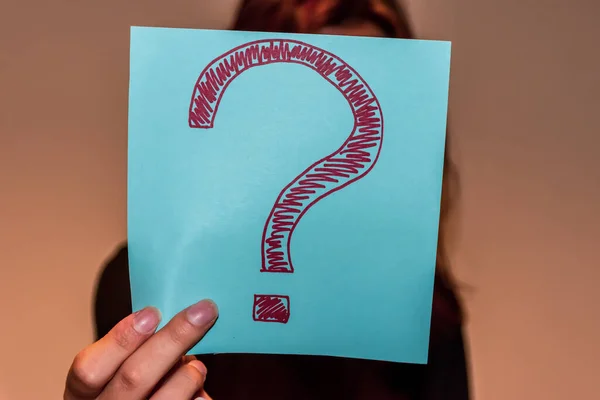 A close-up shot of an unrecognizable young Caucasian redhead woman\'s hand holding a light blue paper sheet with a hand-drawn question mark and hiding her face behind it