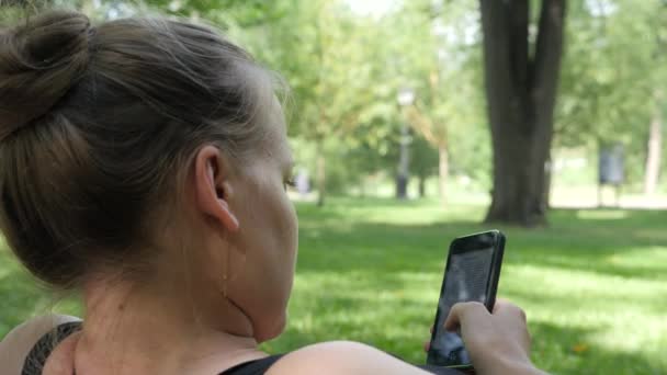 Молодая женщина лежит на лужайке и читает книгу по телефону. Парк в солнечный кипящий день — стоковое видео