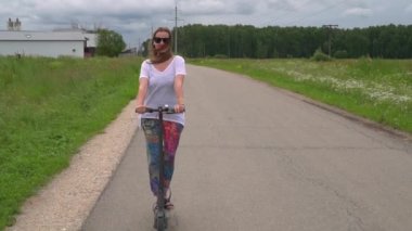 Gülümseyen kadın itme scooter ile kırsal kesimde asfalt yolda gidiyor. Ağır çekim vurdu