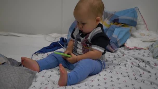 Il bambino si siede in un letto e guarda il cellulare nelle sue mani. Colpo palmare — Video Stock
