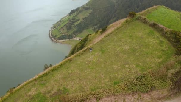 Hiker walks on green meadow at the edge of caldera above lake. Aerial of Sete Cidades volcano, San Miguel, Azores — 图库视频影像