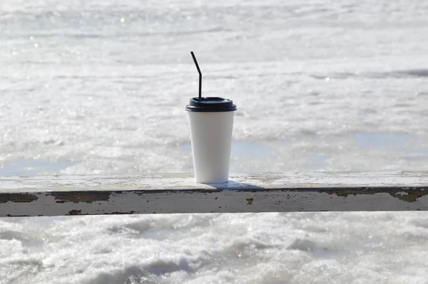 Coffee in the plastic cup standing on wooden bench against the background of melting sea in early spring