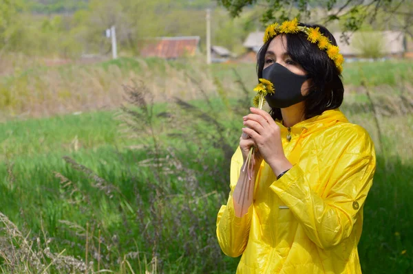 Brunette woman in yellow jacket with wreath of dandelions and medical black mask sniffing bouquet of dandelions. looking away. coronavirus pandemic concept.