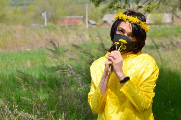 Brunette woman in yellow jacket with wreath of dandelions and medical black mask sniffing bouquet of dandelions. looking at camera.coronavirus pandemic concept.