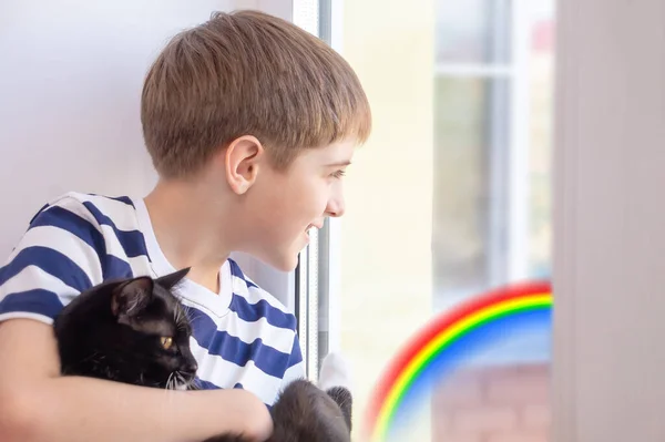 A boy in a t-shirt with a black cat sits on the windowsill and looks out the window. The image of the rainbow on the window. Flash mob of the society for self-isolation from quarantine coronavirus.