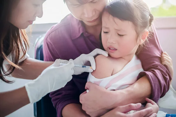 Маленький ребенок с инъекцией, крупным планом Доктор вводит вакцинацию к руке азиатской маленькой девочки, с отцом обнимает ребенка, за то, что он не извивается во время инъекции вакцины — стоковое фото