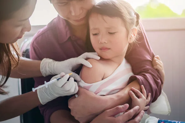 Маленький ребенок с инъекцией, крупным планом Доктор вводит вакцинацию к руке азиатской маленькой девочки, с отцом обнимает ребенка, за то, что он не извивается во время инъекции вакцины — стоковое фото
