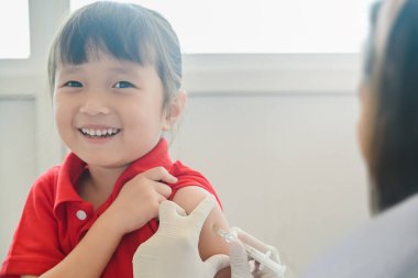 Küçük Asyalı çocuk enjeksiyon oluyor, Doktor küçük kızın gülümseyen yüzüne aşı enjekte ediyor ve kameraya bakıyor, aşı aşısı sağlık ve tıbbi konsept için.