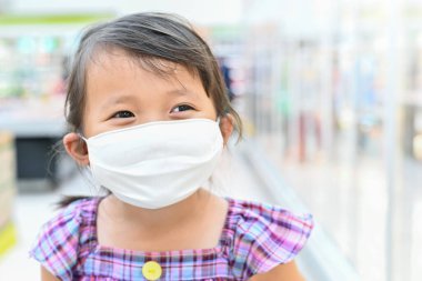 Küçük kızın kumaş maskesi var. Çocuk süpermarketten sonra annesiyle süpermarkete gittiğinde kendini Coronavirus 'tan koruyor. Salgından sonra açık hava aktivitesi için burnuna maske takmış bir çocuk..