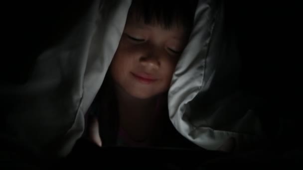 Küçük kız gece yatağında tablette video izliyor ekrandan yansıyan ışıklar, bağımlılık ve çizgi film kavramlarıyla oyun oynayan çocuklar. — Stok video