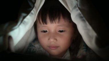 Küçük kız gece yatağında cep telefonuyla video izliyor ekrandan yansıyan ışıklar, bağımlılık ve çizgi film kavramlarıyla oyun oynayan çocuklar.