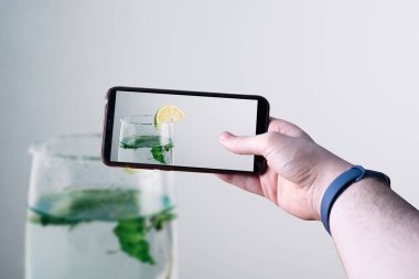 Akıllı telefonu kaldırıp ahşap bir masada limonlu mojitonun fotoğrafını çeken eller. Mesaj için yer var. Seçici odak