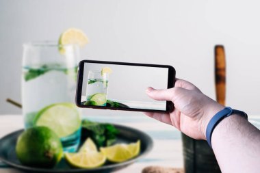 Akıllı telefonu kaldırıp ahşap bir masada limonlu mojitonun fotoğrafını çeken eller. Mesaj için yer var. Seçici odak