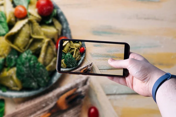 Akıllı telefonu kaldırıp ahşap masada içinde malzemeler olan taze bir makarnanın fotoğrafını çeken eller. Mesaj için yer var. Seçici odak.