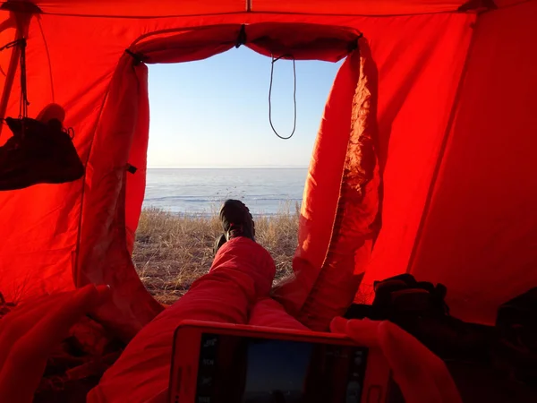 a tourist sits in a red tourist tent, holding a phone, and his feet stick out of the tent\'s exit, he looks at the frozen surface of the lake