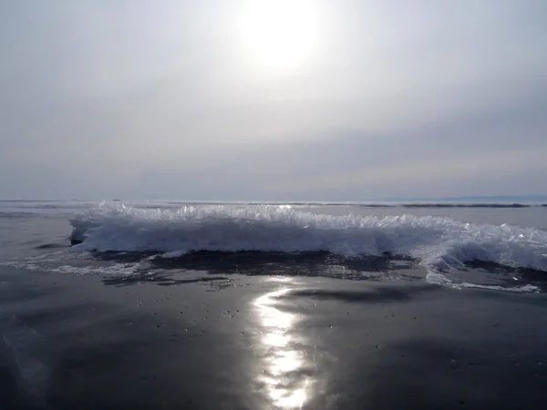 太陽の進路は 白い氷の結晶のある雪が波のクレストの上に上昇するので 氷の暗い表面に反映されます 暗い灰色の空が地平線と合流し — ストック写真