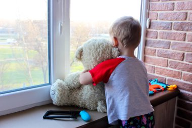 Elinde oyuncak ve ayı olan bir çocuk karantina sırasında pencereden dışarı bakar.
