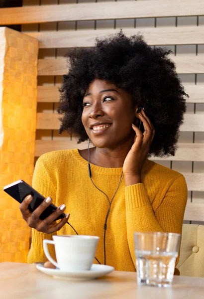 Siyah Afrikalı güzel bir kadın, kafede oturuyor, müzik dinliyor ya da kulaklarında kulaklıklarla cep telefonuyla konuşuyor ve bir fincan kahvenin tadını çıkarıyor..