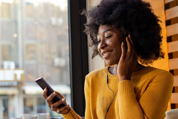 Siyah Afrikalı güzel bir kadın, kafede oturuyor, müzik dinliyor ya da kulaklarında kulaklıklarla cep telefonuyla konuşuyor ve bir fincan kahvenin tadını çıkarıyor..
