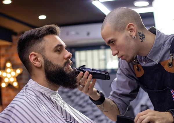 Getting perfect shape, beard styling and cutting with trimmer. Young bearded hipster man getting haircut by hairdresser at barber shop. Advertising and barber shop concept.