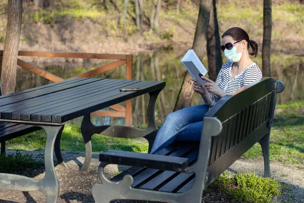 脸上戴着保护膜的女人 坐在湖边公园的长椅上看书 — 图库照片