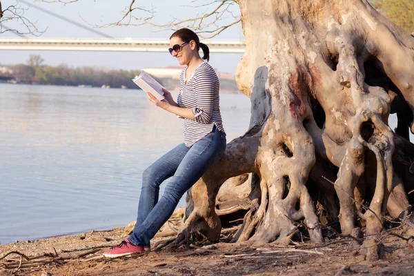 女学生坐在平坦的树根上 在河边看书 — 图库照片