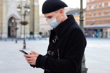 Yüzünde koruyucu maskesi olan genç bir adam boş bir sokakta tek başına buluşmayı beklerken akıllı telefonundan veri topluyor. Covid 19 virüs salgını veya hava kirliliği kavramı.