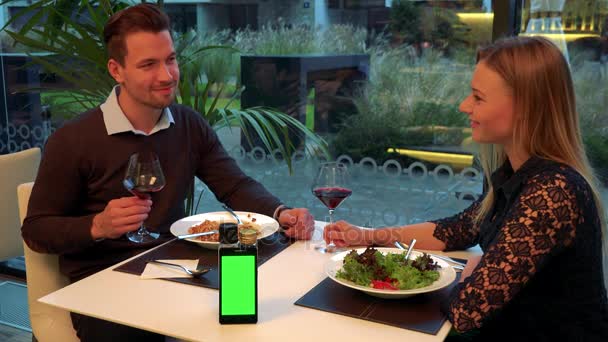 Мужчина и женщина (как молодые, так и привлекательные) сидят за столом в ресторане, улыбаются в камеру и в конце концов обмениваются взглядами - смартфон на столе — стоковое видео
