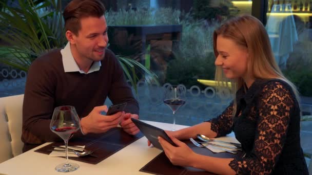 A man and a woman (both young and attractive) sit at a table in a restaurant, the man holds a smartphone, the woman holds a tablet and they show each other something on them — Stock Video