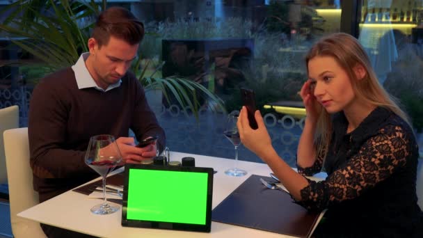 Un uomo e una donna si siedono a un tavolo in un ristorante, lui scrive su uno smartphone, lei controlla il trucco e i capelli in un riflesso del suo smartphone. — Video Stock