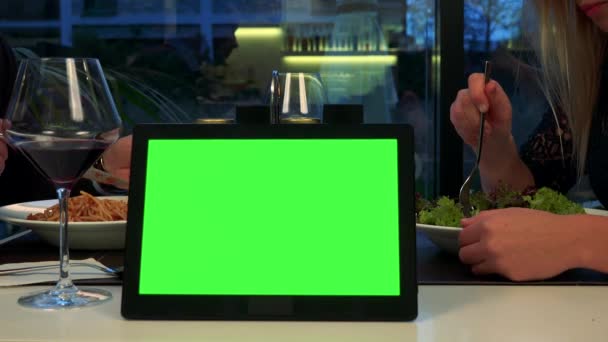 Een man en een vrouw zitten aan een tafel en eten, een tablet met een groen scherm op de voorgrond — Stockvideo