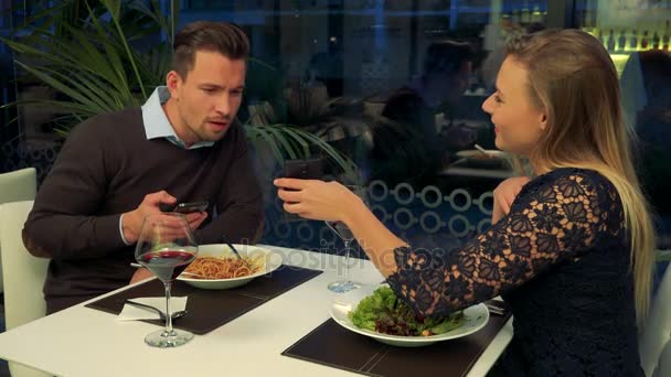 Een man en een vrouw (zowel jong en mooi) zitten aan een tafel in een restaurant, hij heeft zijn smartphone, ze toont hem iets op haar smartphone — Stockvideo