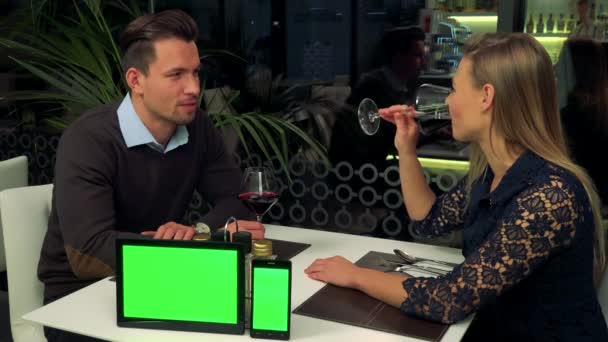 A man and a woman (both young and attractive) sit at a table in a restaurant and talk, a tablet and a smartphone (both green screens) sit on a table, closer to the camera — Stock Video