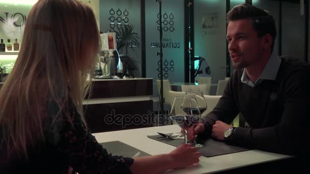 Een jonge, knappe man en een vrouw zitten aan een tafel in een restaurant, praten, dan clink glazen wijn en drinken — Stockvideo