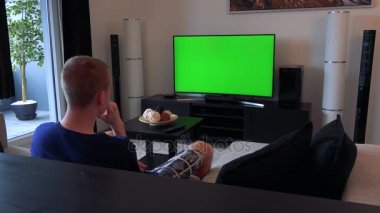 Bir adam rahat bir oturma odasında yeşil bir ekran bir Tv izliyor, sonunda uzakta görünüyor