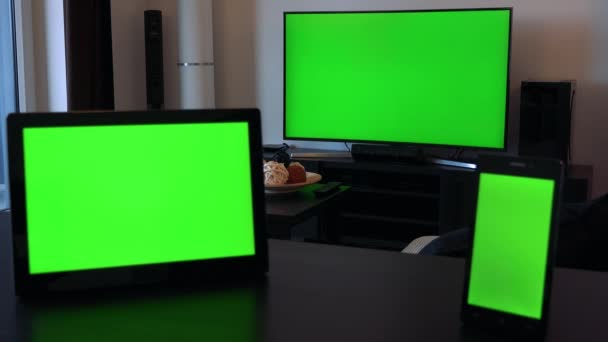 Um tablet (horizontal) e um smartphone (vertical) em uma mesa, uma TV em segundo plano - todos os três com telas verdes — Vídeo de Stock